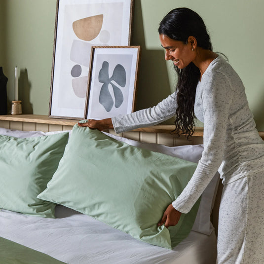 Woman arranging green pillows on a bed in a bedroom setting.