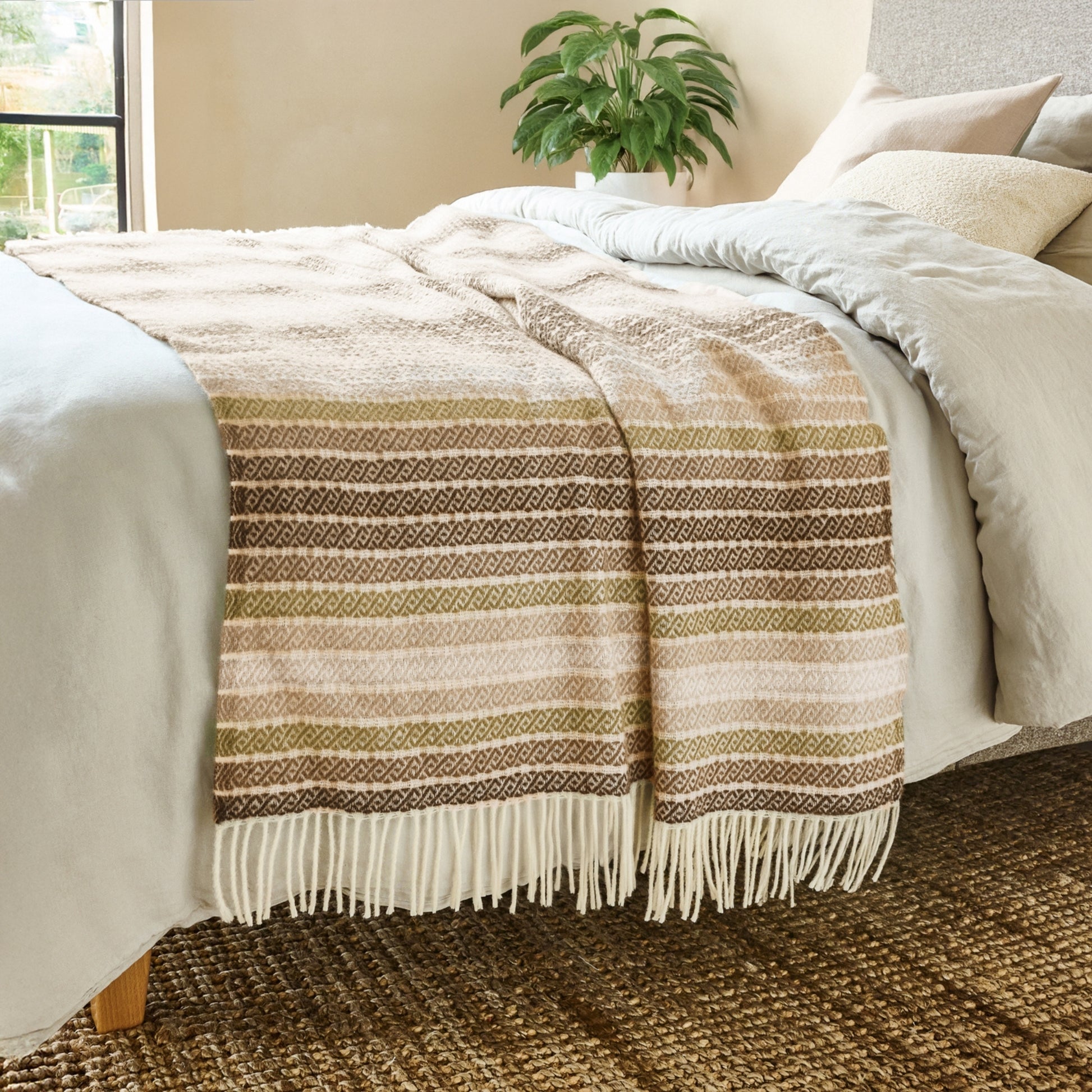 The Athena Wool Throw in the color Clay is a Greek-derived woven pattern, shown draped over the end of a bed with white fringe.