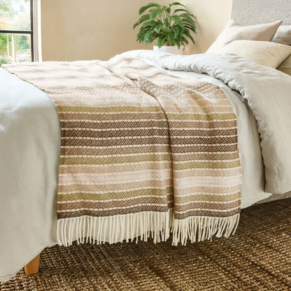 The Athena Wool Throw in the color Clay is a Greek-derived woven pattern, shown draped over the end of a bed with white fringe.