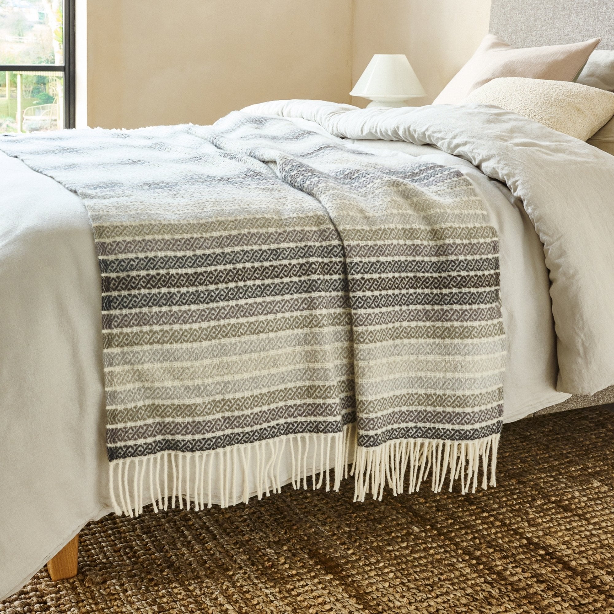 The Athena Wool Throw in the color Stone is a Greek-derived woven pattern, shown draped over the end of a bed with white fringe.