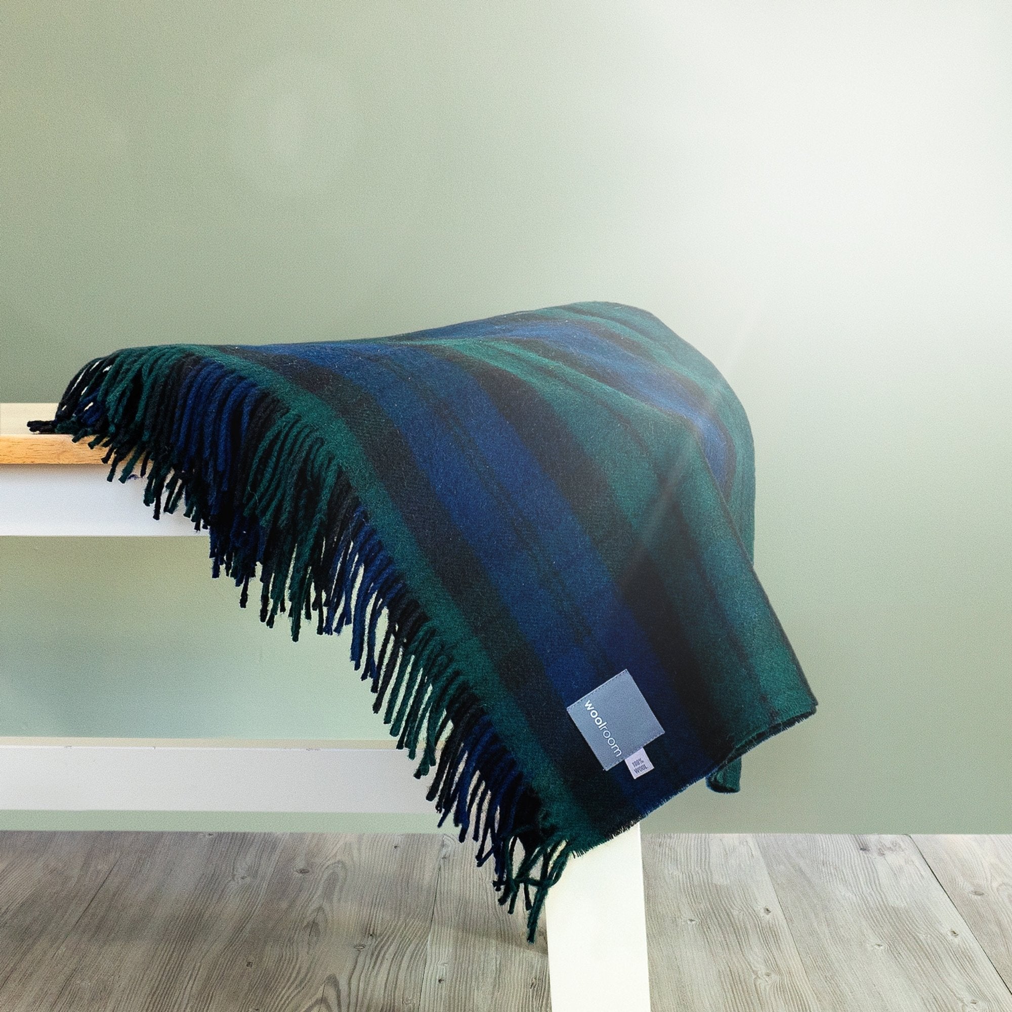 The Blackwatch Tartan Throw in Blue and Green is folded and hanging over the edge of a bench showing the fringe. 