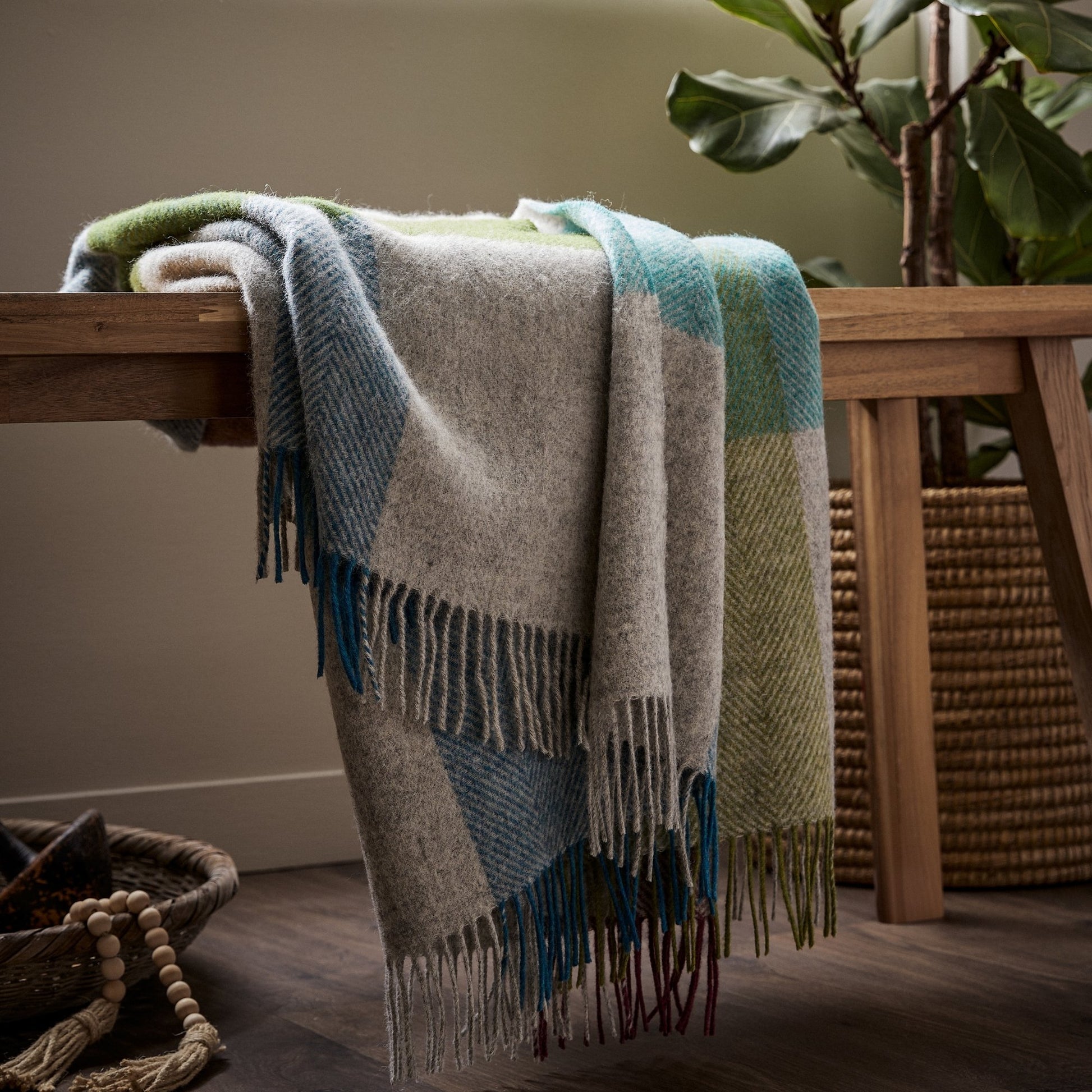 The Harland Shetland Wool Throw in Heather is a mix of oatmeal, blues, greens and purple, draped over a bench with fringe. 