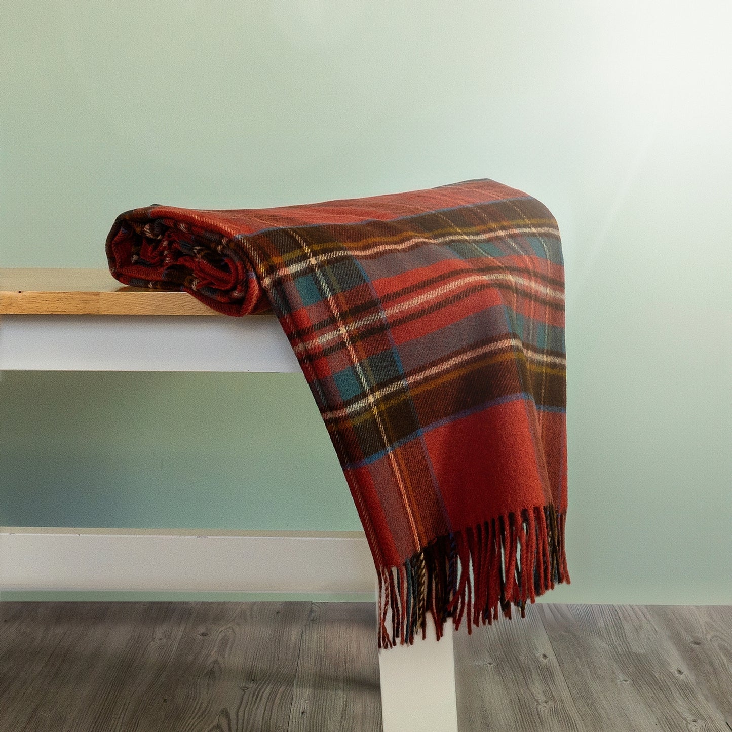 The Antique Royal Stewart Tartan Throw in Orange and Brown is rolled and hanging over the edge of a bench showing the fringe. 