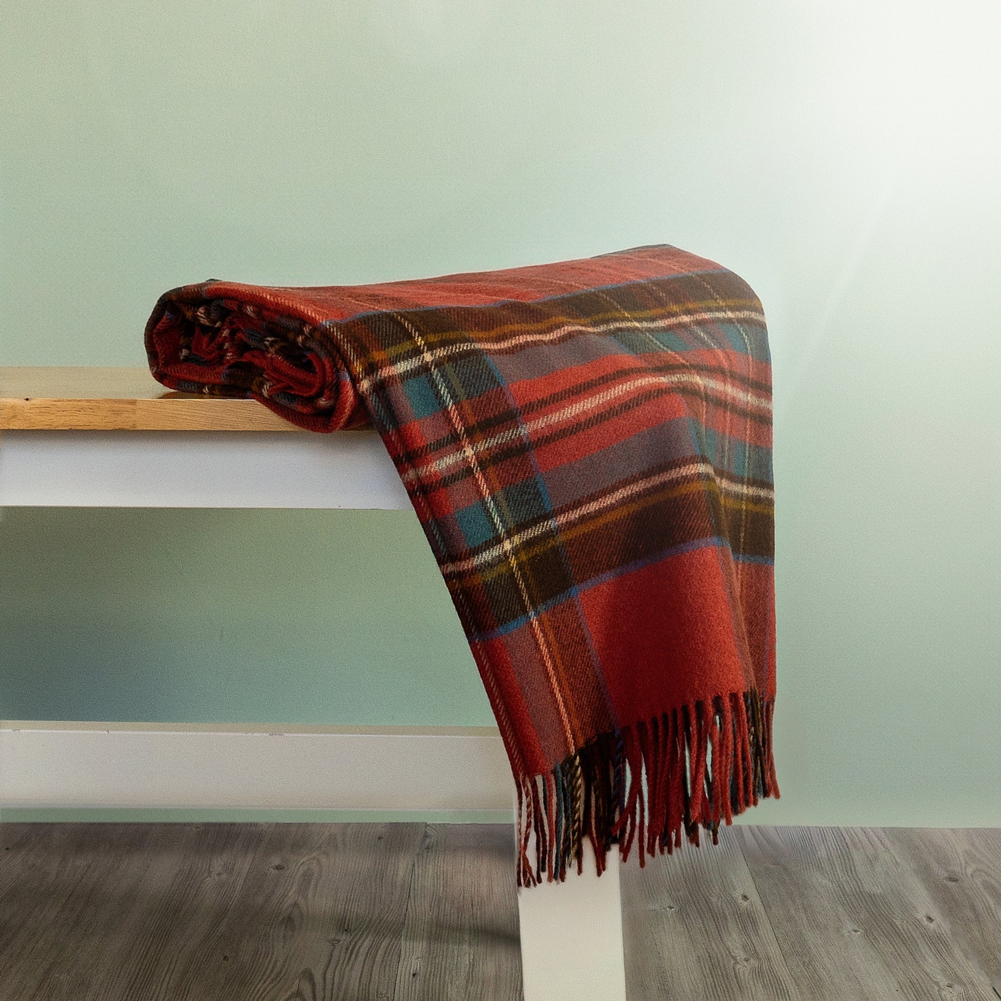 The Antique Royal Stewart Tartan Throw in Orange and Brown is rolled and hanging over the edge of a bench showing the fringe. 