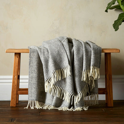 The super soft Greta Herringbone Wool Blanket in Grey draped over a bench with white fringe. 
