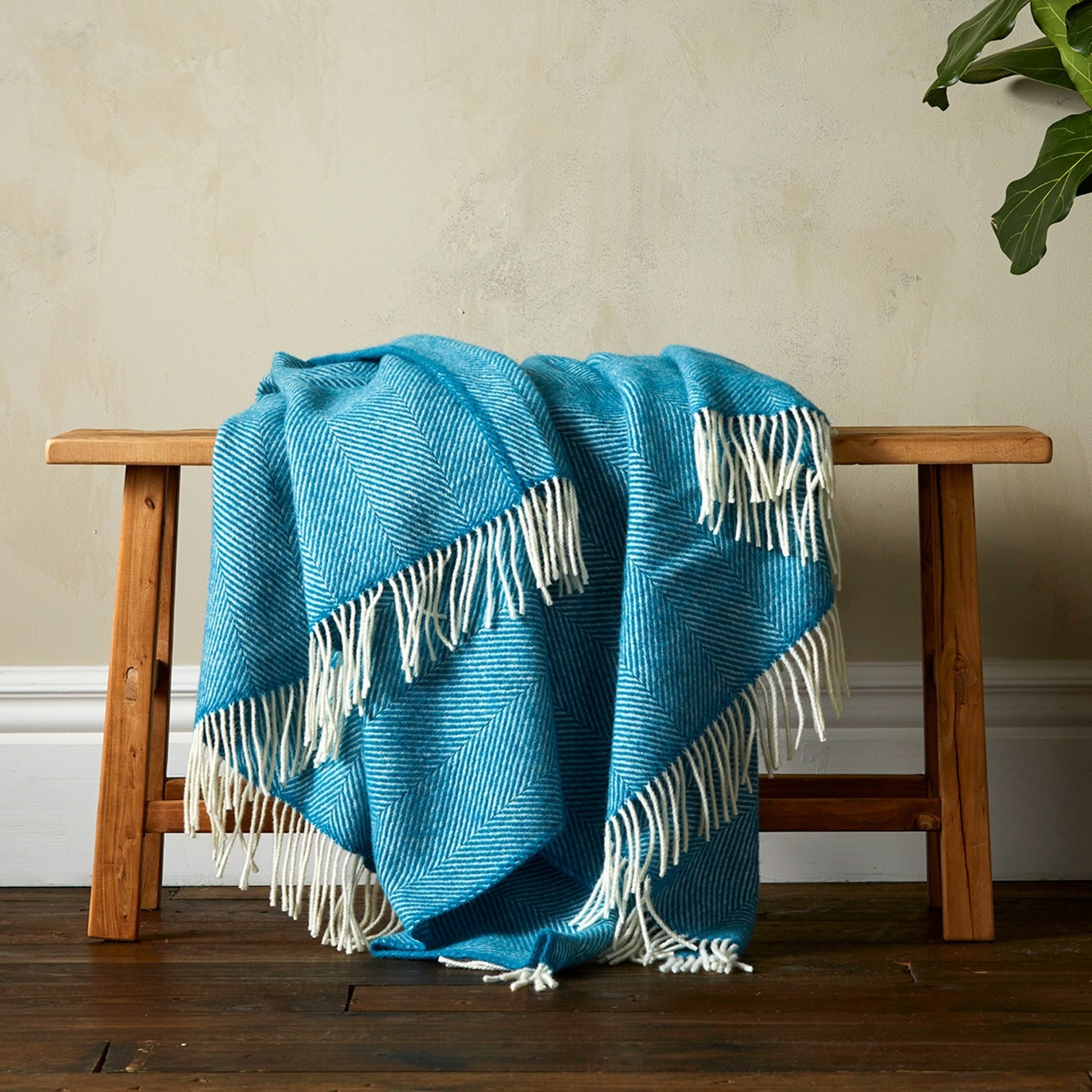 The super soft Greta Herringbone Wool Blanket in Teal draped over a bench with white fringe. 