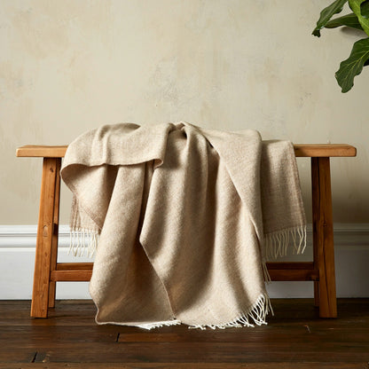 The Woolroom Lilly Herringbone Pure New Wool Throw in Beige has a finer pattern detail and is draped over a bench with fringe.