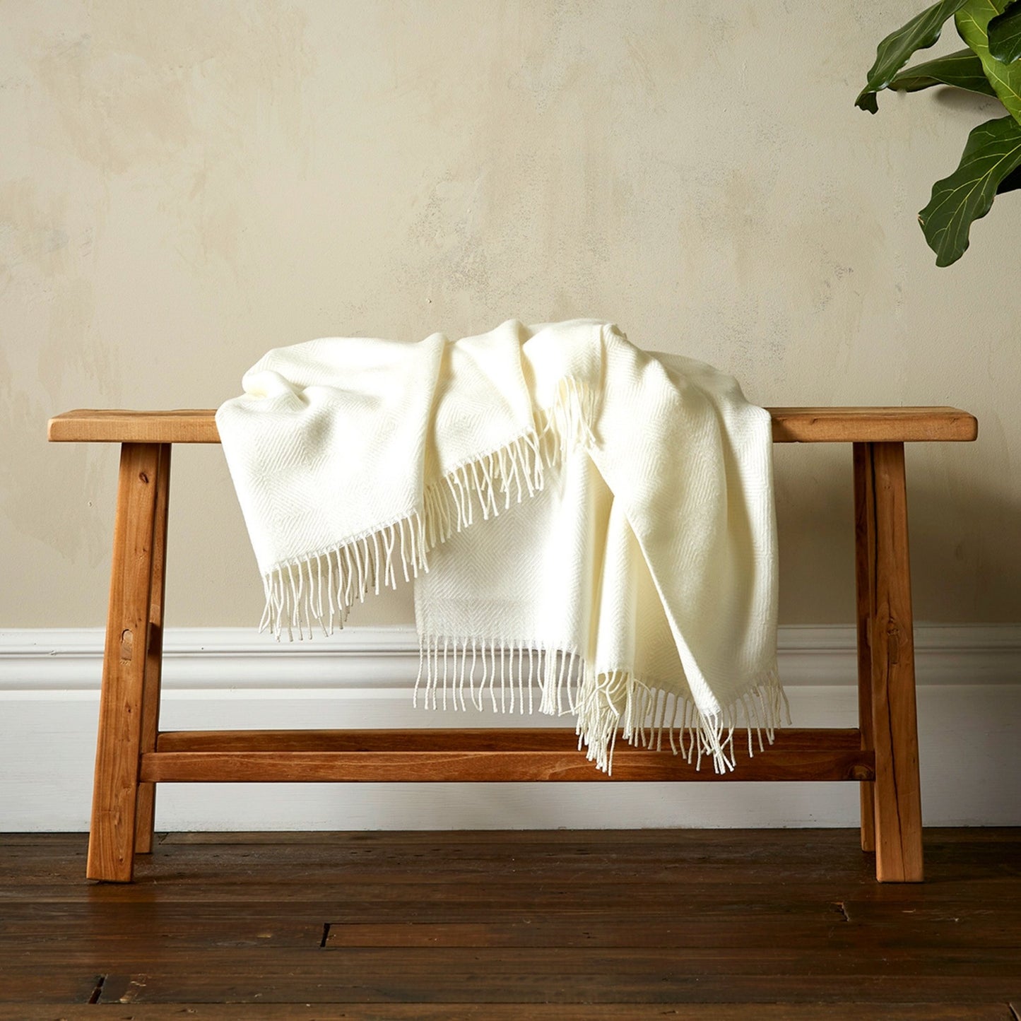 The Woolroom Diamond Twill Throw is a pure white fine weave with white fringe, draped over a bench. 