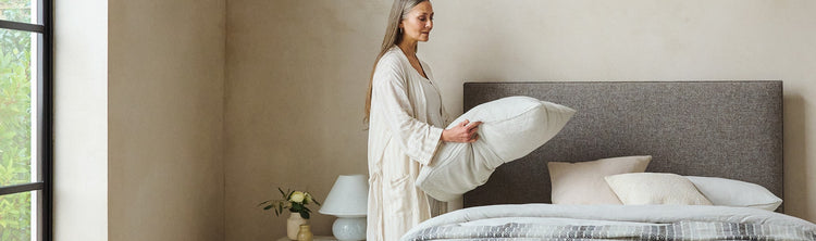 Woman plumping a pillow on a bed dressed in pale linen and a grey-and-cream striped wool throw.
