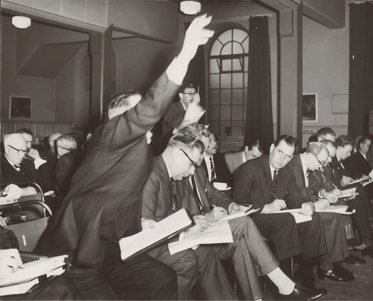 Black and white photograph of a seated audience or meeting room, with several men in suits writing in notebooks and one man raising his hand. The scene is indoors with windows, curtains, and architectural details visible.