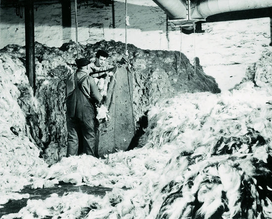 Black and white historical photograph showing two men standing in a snowy outdoor setting, holding a large metal panel or door in place amidst snow and industrial structures above.