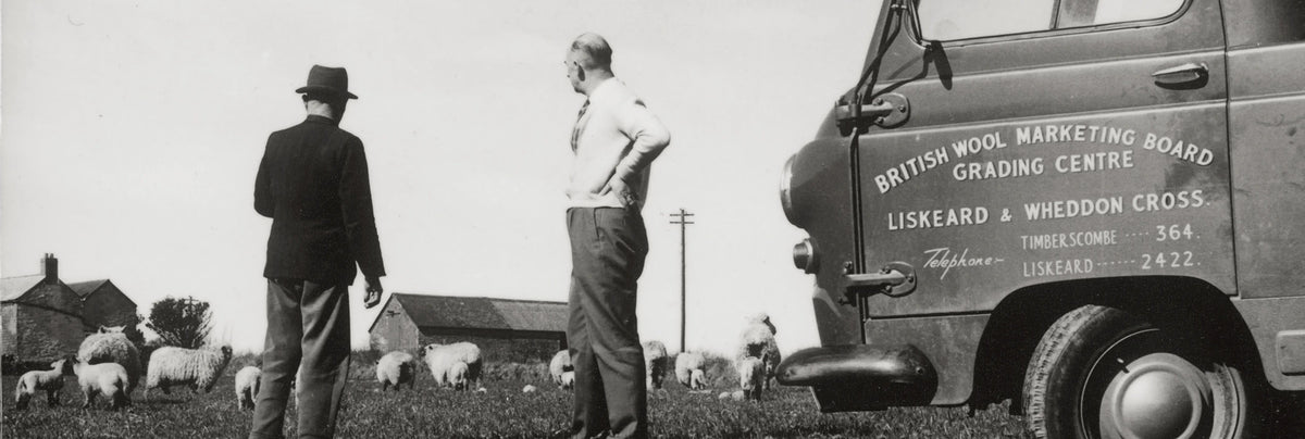 Black and white photograph showing two men standing beside a vintage truck in a rural field with sheep in the background. The truck has visible text reading 'BRITISH WOOL MARKETING BOARD GRADE CENTRE LISKEDWE AND WEDDON CROSS'.