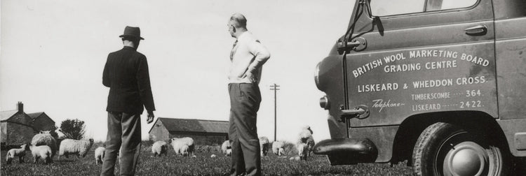 Black and white photograph showing two men standing beside a vintage truck in a rural field with sheep in the background. The truck has visible text reading 'BRITISH WOOL MARKETING BOARD GRADE CENTRE LISKEDWE AND WEDDON CROSS'.
