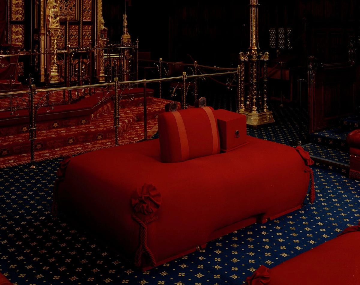 A red ceremonial throne or sofa-like furniture piece in a dark ornate interior, with blue patterned carpet and gold-toned architectural details in the background.