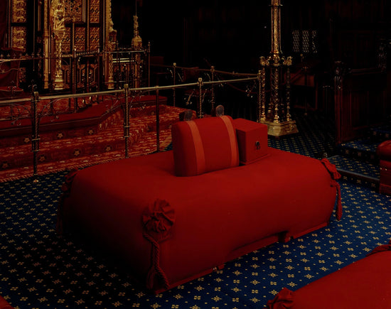 A red ceremonial throne or sofa-like furniture piece in a dark ornate interior, with blue patterned carpet and gold-toned architectural details in the background.