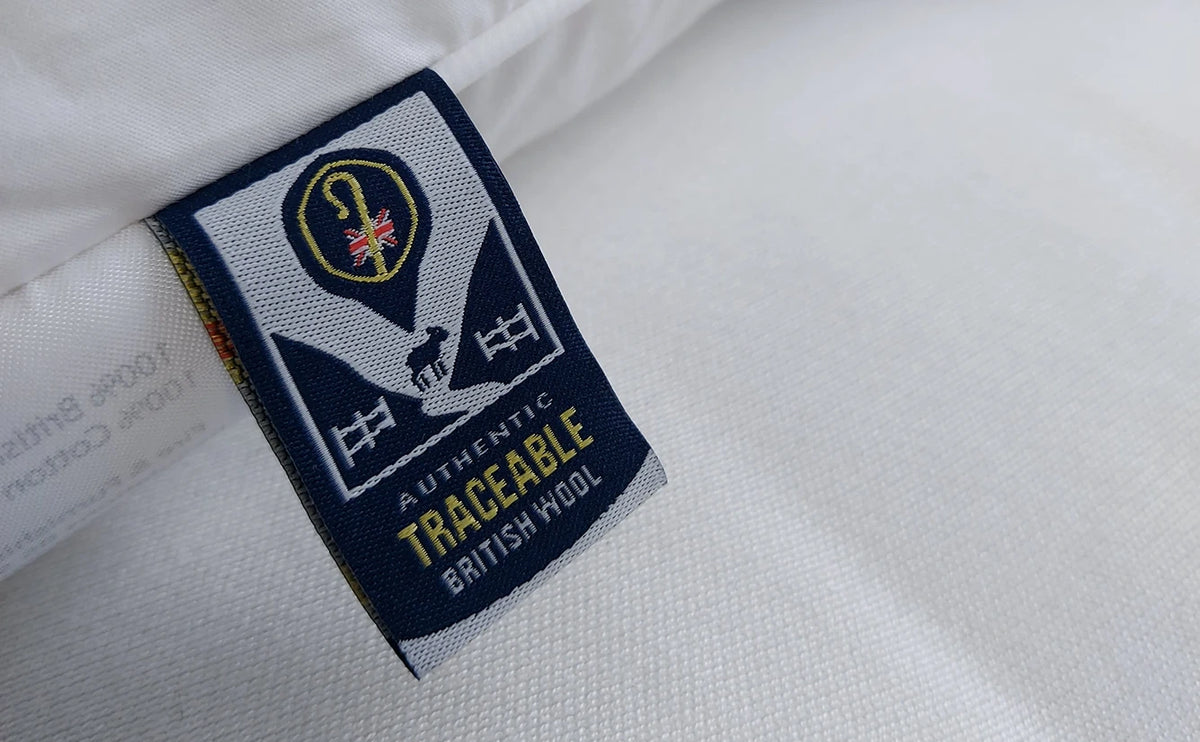 Close-up of a white woven textile with a sewn fabric label reading 'AUTHENTIC TRACEABLE BRITISH WOOL' and a crest-style logo, showing the material texture and label details.