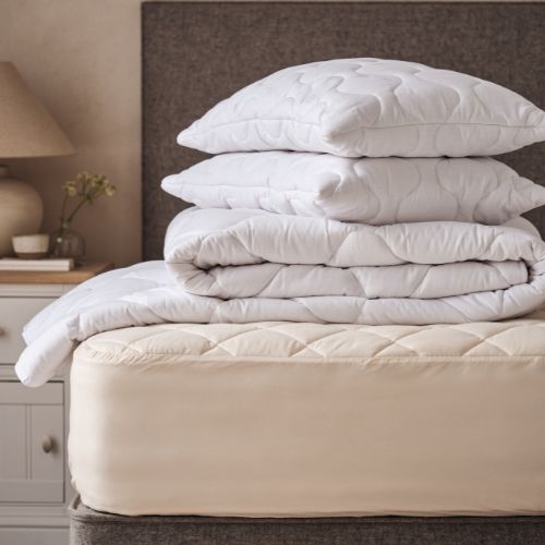 A blonde lady sitting on a mattress, with a pile of Woolroom bedding, including a duvet, pillows and mattress protector
