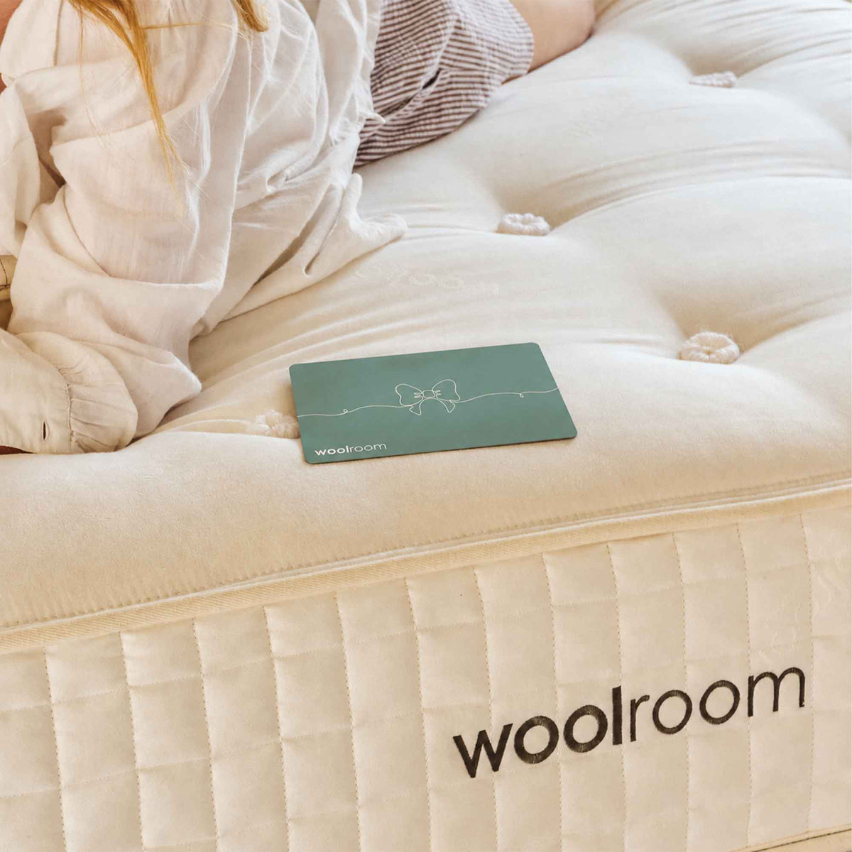 A close-up of a cream tufted mattress with quilted side panels, showing the Woolroom brand name. A person in a white shirt is partially visible lying on the mattress, and a green gift card with the Woolroom logo rests on the mattress surface.