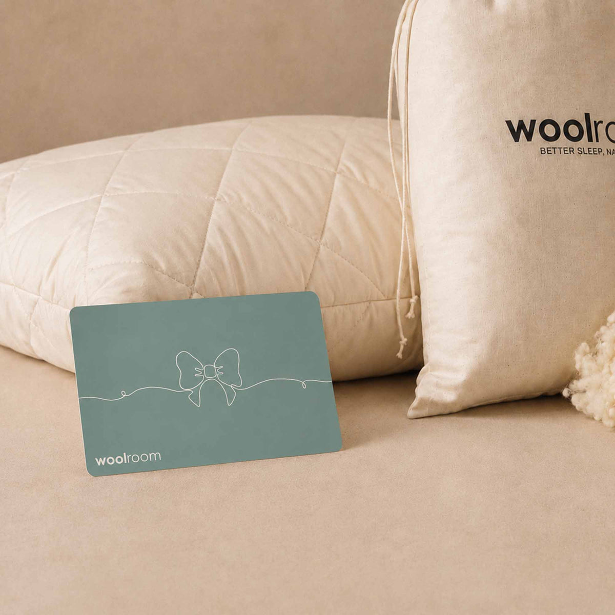 A teal Woolroom gift card with a bow logo lies on a beige surface in front of cream-colored pillows, including one with the Woolroom branding printed on it.