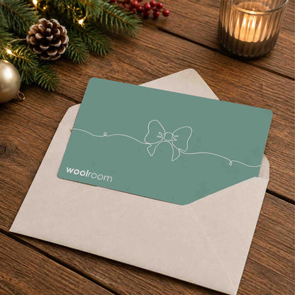 A teal greeting card with a white bow line drawing and the Woolroom logo, placed on top of a kraft paper envelope on a wooden table with holiday decorations and a lit candle in the background.