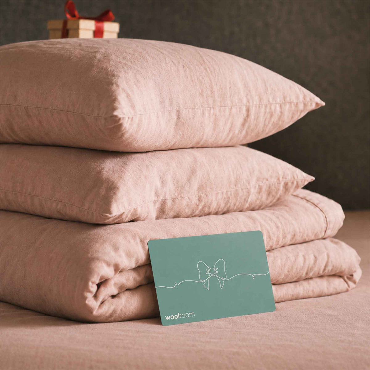 A stack of blush pink bedding, including two pillows and a folded duvet cover, displayed on a bed with a teal wooloom gift card in the foreground.
