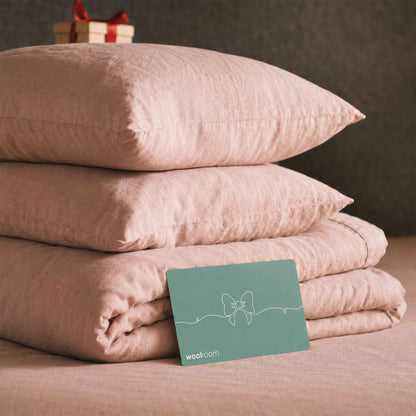 A stack of blush pink bedding, including two pillows and a folded duvet cover, displayed on a bed with a teal wooloom gift card in the foreground.