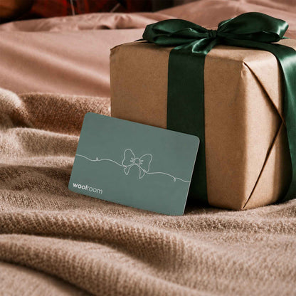A Woolroom gift card with a sage green background and white bow line-art sits in front of a brown kraft paper gift box tied with a dark green ribbon, styled on a beige knit blanket.
