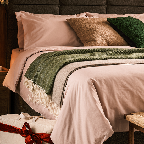 Neatly made bed with pink bedding and green throw, accompanied by a wrapped gift.