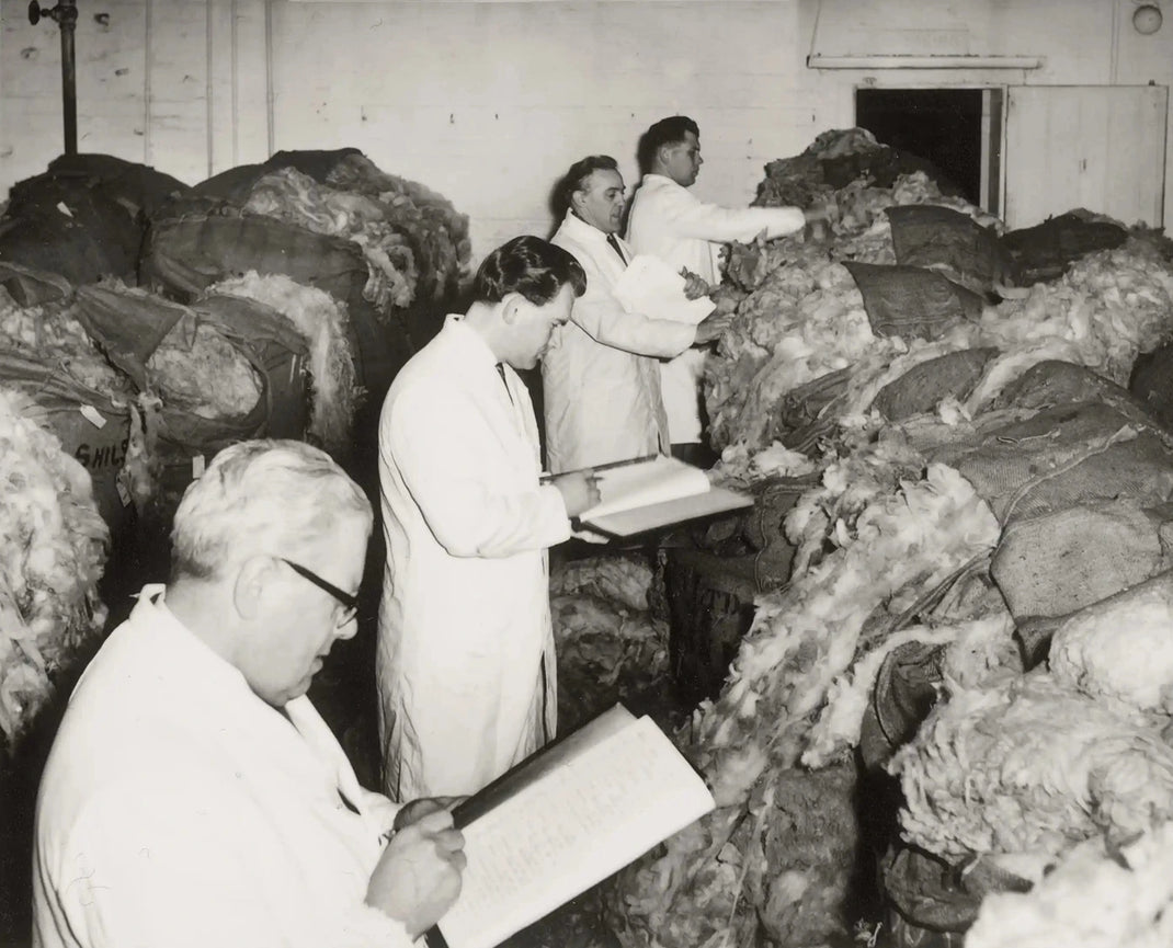 Black-and-white archival photo of men in white coats inspecting large packed wool bales in a warehouse.
